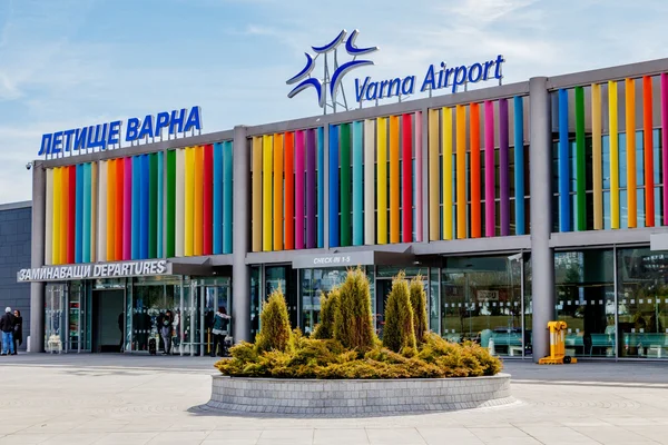 Varna Airport terminal building with colorful vertical stripes on the facade and landscaping in front under a blue sky.