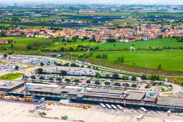 Aerial view of Verona Villafranca Airport with runways, terminal buildings, and parked aircraft, surrounded by green farmland and a residential town in the background.