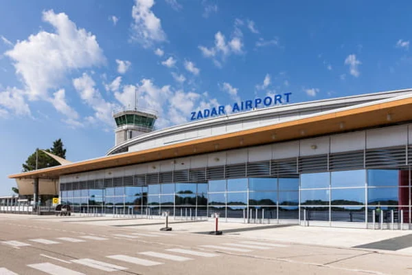 Modern Zadar Airport terminal building with control tower, large glass windows, and paved forecourt under blue sky with white clouds.