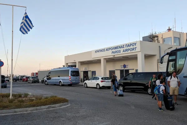 Exterior view of Paros National Airport terminal building with parked vehicles, flags, and passengers during daytime.
