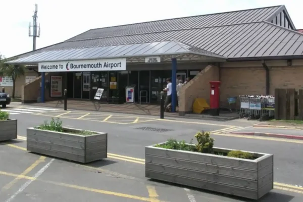 Widok zewnętrzny terminalu Lotniska Bournemouth z nowoczesną architekturą, drzwiami wejściowymi i betonowymi doniczkami na terenie przed budynkiem.