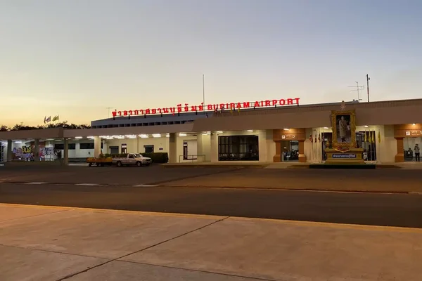 Терминал аэропорта Бури-Рам, освещенный в сумерках с припаркованными автомобилями.