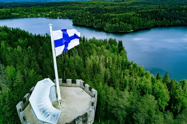 Une vue aérienne de la tour d'observation Aulanko à Hämeenlinna, en Finlande, présentant un majestueux drapeau finlandais sur fond de forêts verdoyantes et de lacs sereins.