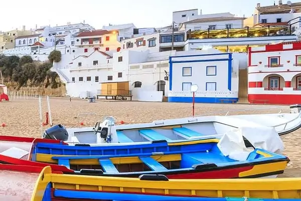 In the foreground the colourful boats on a beach, in the background the white houses with the colourful boride of Faro in Portugal.