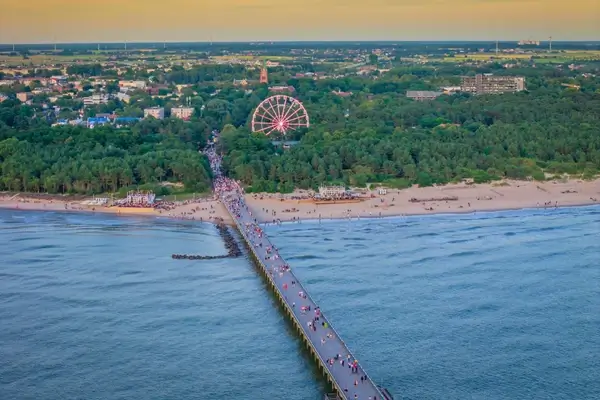 Une vue aérienne de la plage de Palanga au coucher du soleil, avec un quai animé menant au rivage, une grande roue vibrante au milieu d'une verdure luxuriante, et la vaste mer Baltique au premier plan.