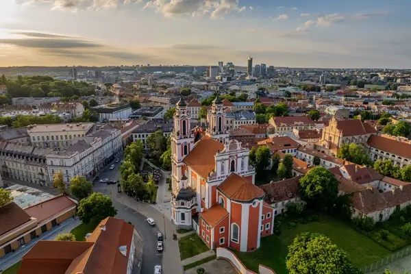 Une vue aérienne de la vieille ville de Vilnius au coucher du soleil, mettant en valeur l'architecture baroque saisissante de l'église Sainte-Catherine, entourée de bâtiments historiques et de verdure luxuriante.
