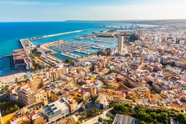 Aerial view of Alicante, Spain, showing the Mediterranean coastline, marina filled with boats, long harbor breakwater, and dense colorful buildings along the shore.
