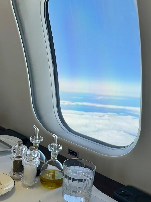 Blick aus dem Kabinenfenster eines Privatjets auf die Wolken, daneben ein gedeckter Esstisch mit Gewürzen und Wasserglas.