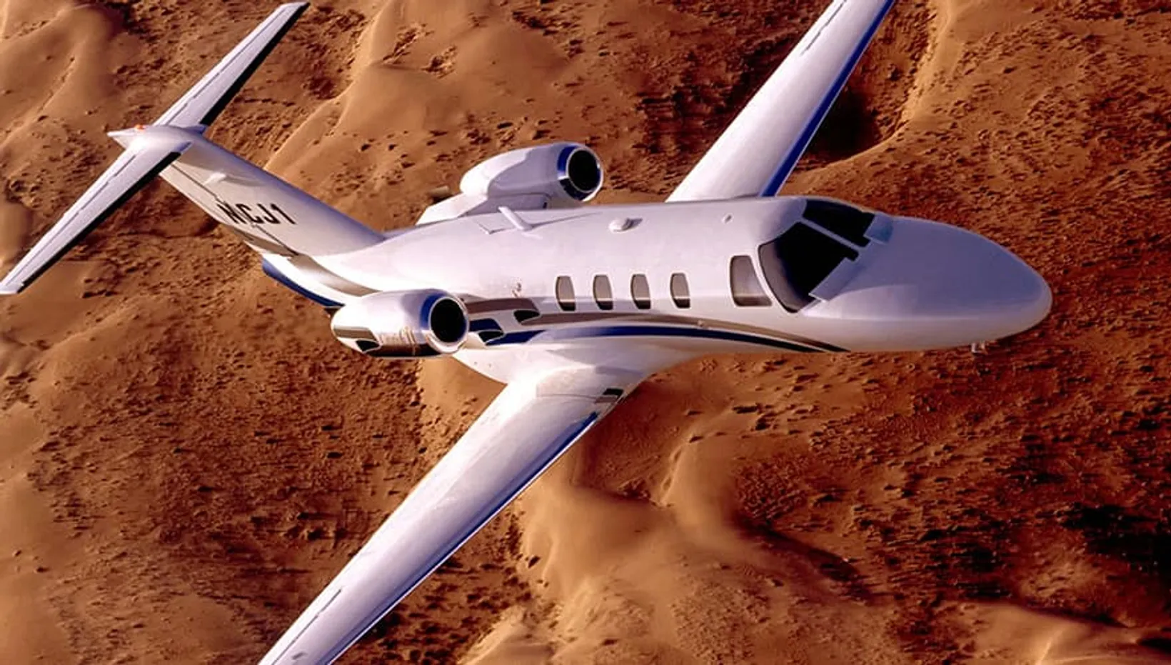 Cessna Citation CJ1 private jet in flight over desert terrain, aerial side view showing twin engines and blue striping.