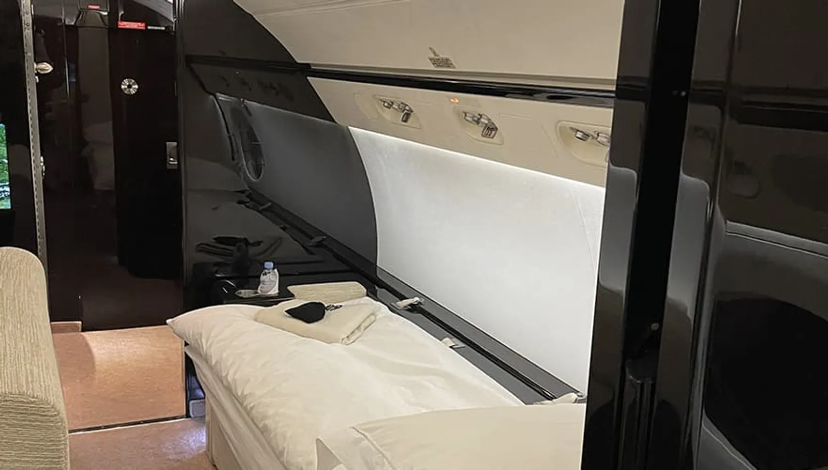 Bombardier Challenger 650 cabin interior showing a cream-colored bed with white bedding, black furniture, and beige wall p...