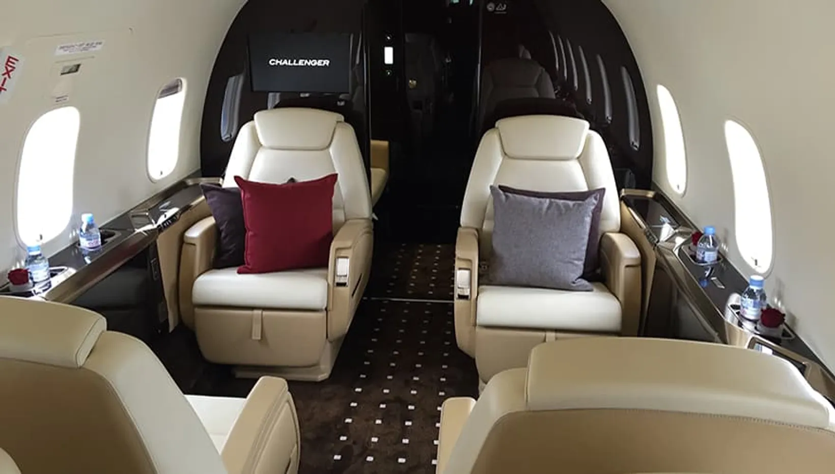 Interior cabin view of Bombardier Challenger 350 jet with cream leather seats, decorative pillows, windows, wooden accents...