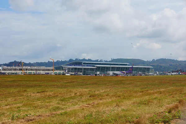 Phuket International Airport terminal and aircraft stands viewed across the airfield, with wooded hills behind