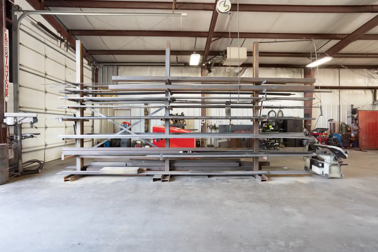 Heavy-duty fabrication shop rack with stacked steel beams and tubing, chop saw, drill press, “FIRE EXTINGUISHER” sign visible.