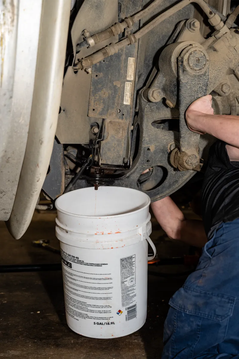 Brake service with fluid draining from a wheel hub into a white 5-gallon bucket marked SAE 5W-40.