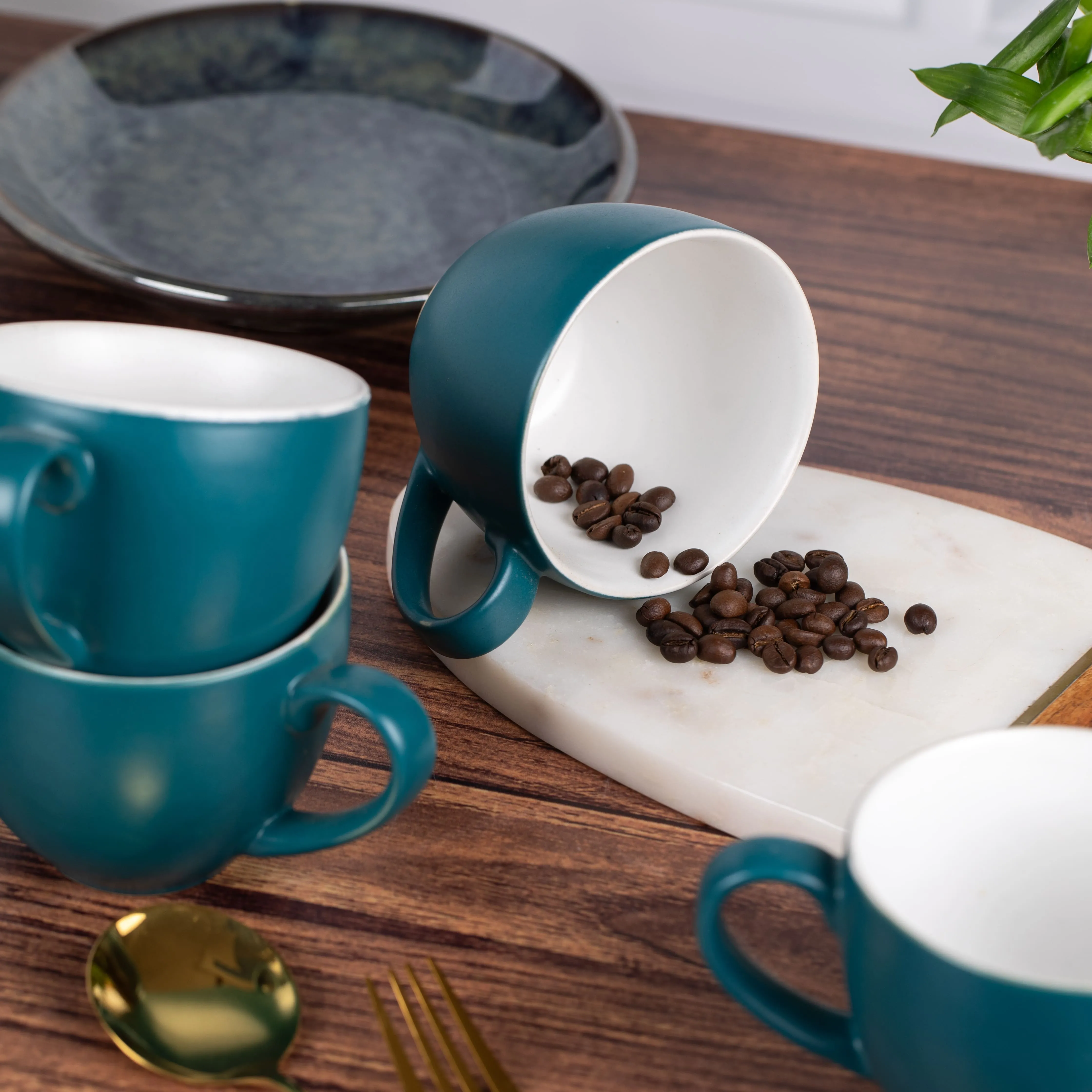 Teal ceramic coffee cups on a wooden table, one tipped over spilling coffee beans onto a white marble board.