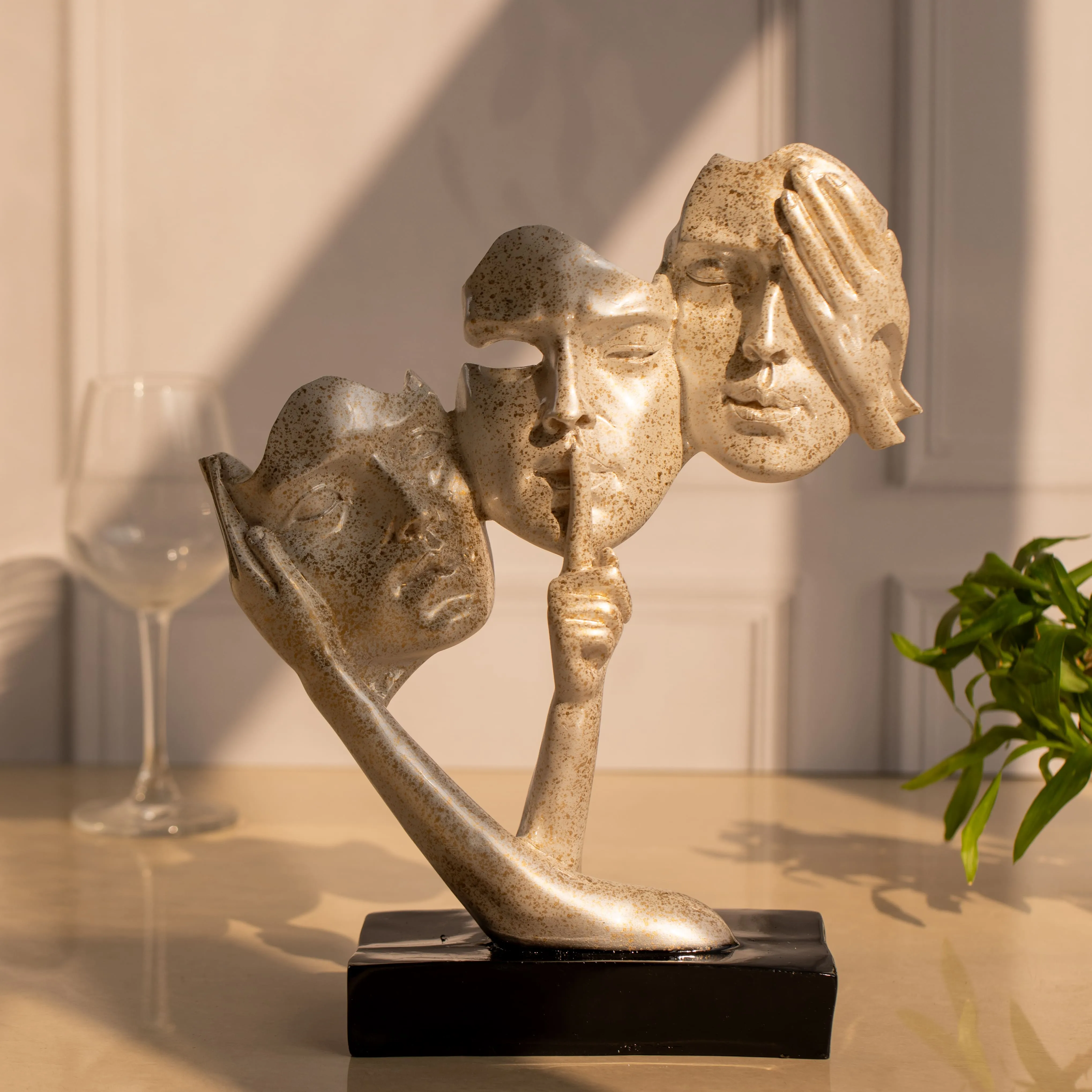 Abstract sculpture with three speckled beige faces and two hands on a black base, set on a table with a wine glass and green plant in soft sunlight.