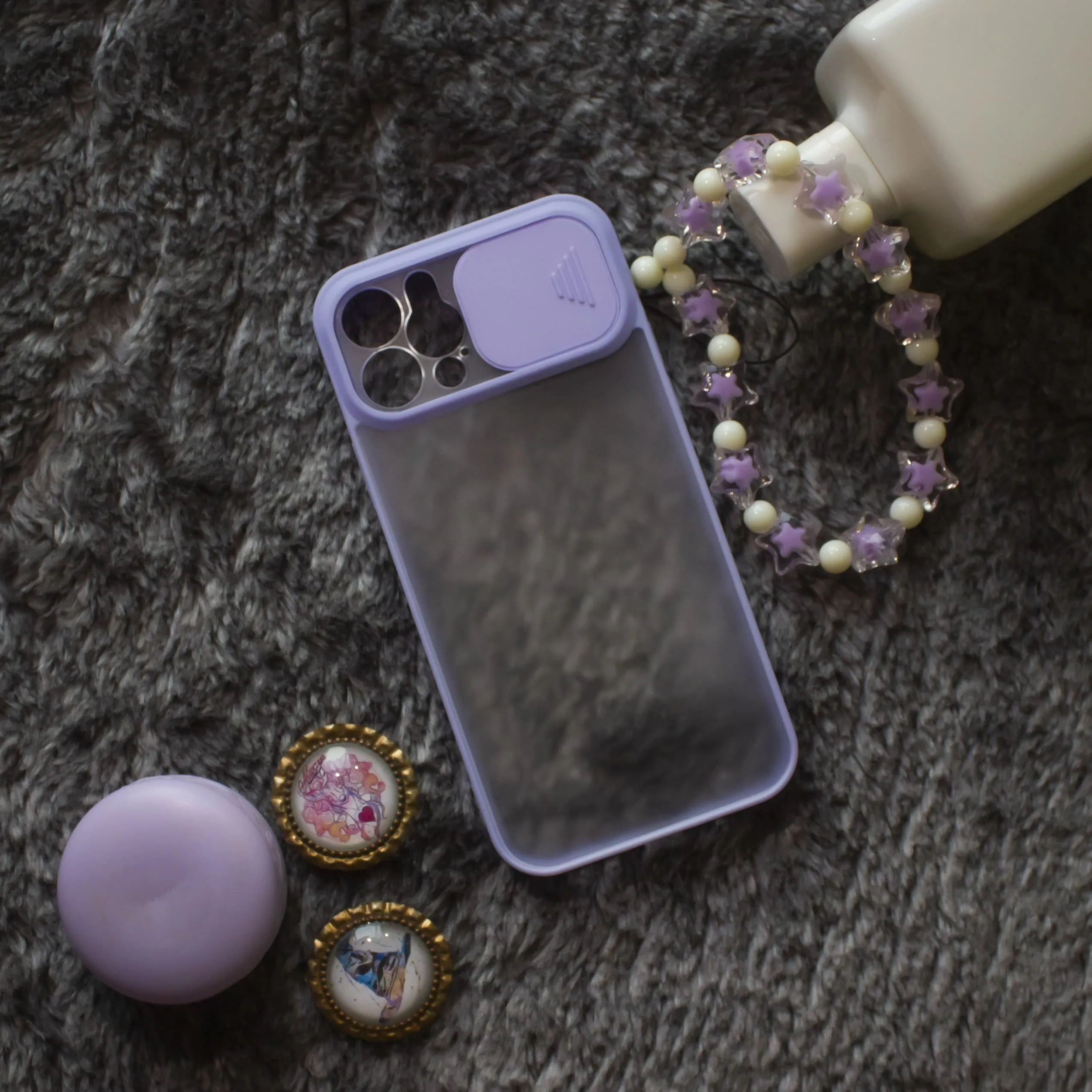 A translucent phone case with purple edges next to a purple circular container, two decorative pins with colorful designs, a beaded bracelet with purple stars, and a white bottle on a dark textured surface.