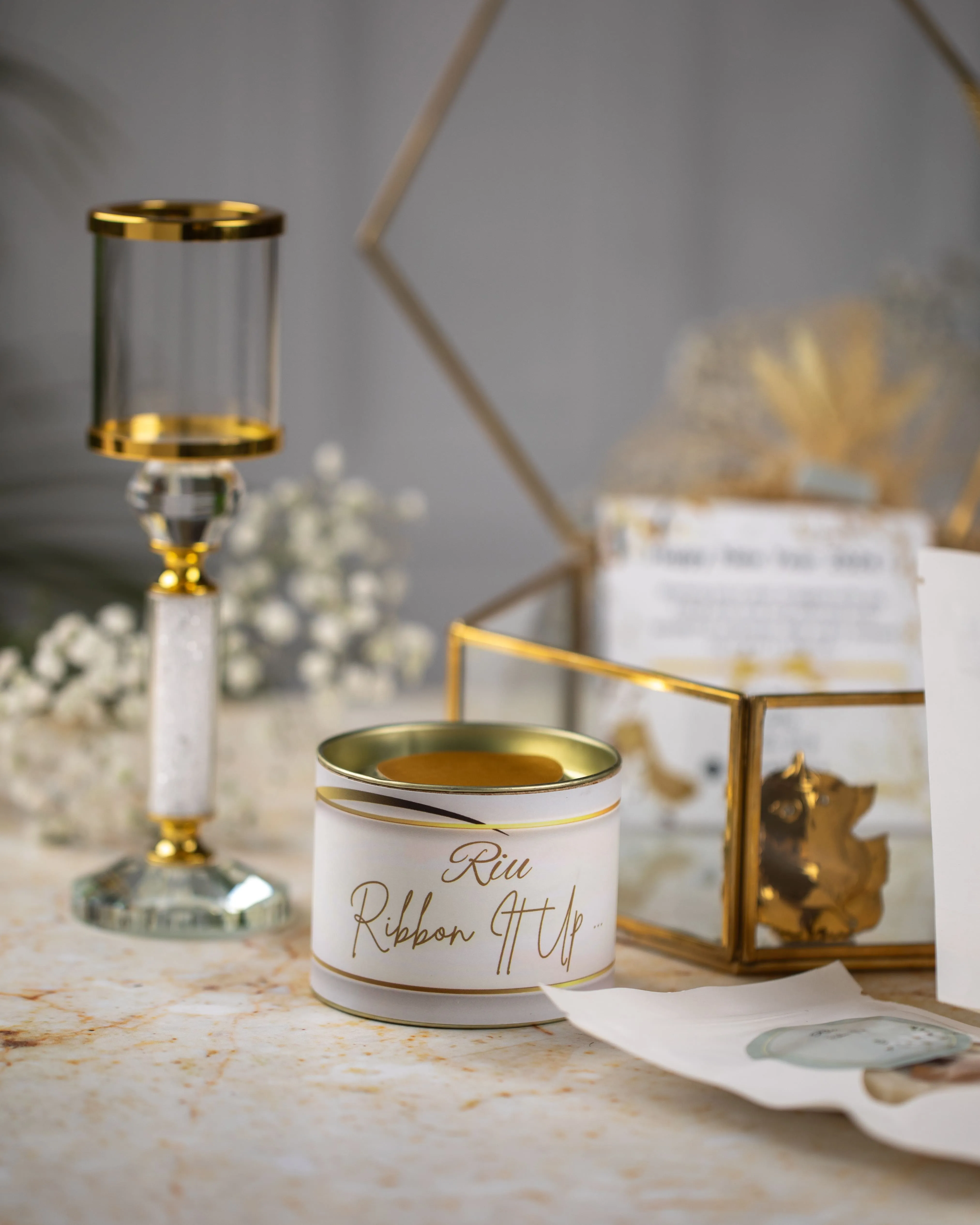 Decorative table setup with a gold-rimmed glass candlestick, a small round container labeled 'Riu Ribbon' with a gold interior, and a glass box with gold accents holding dried flowers and a card.
