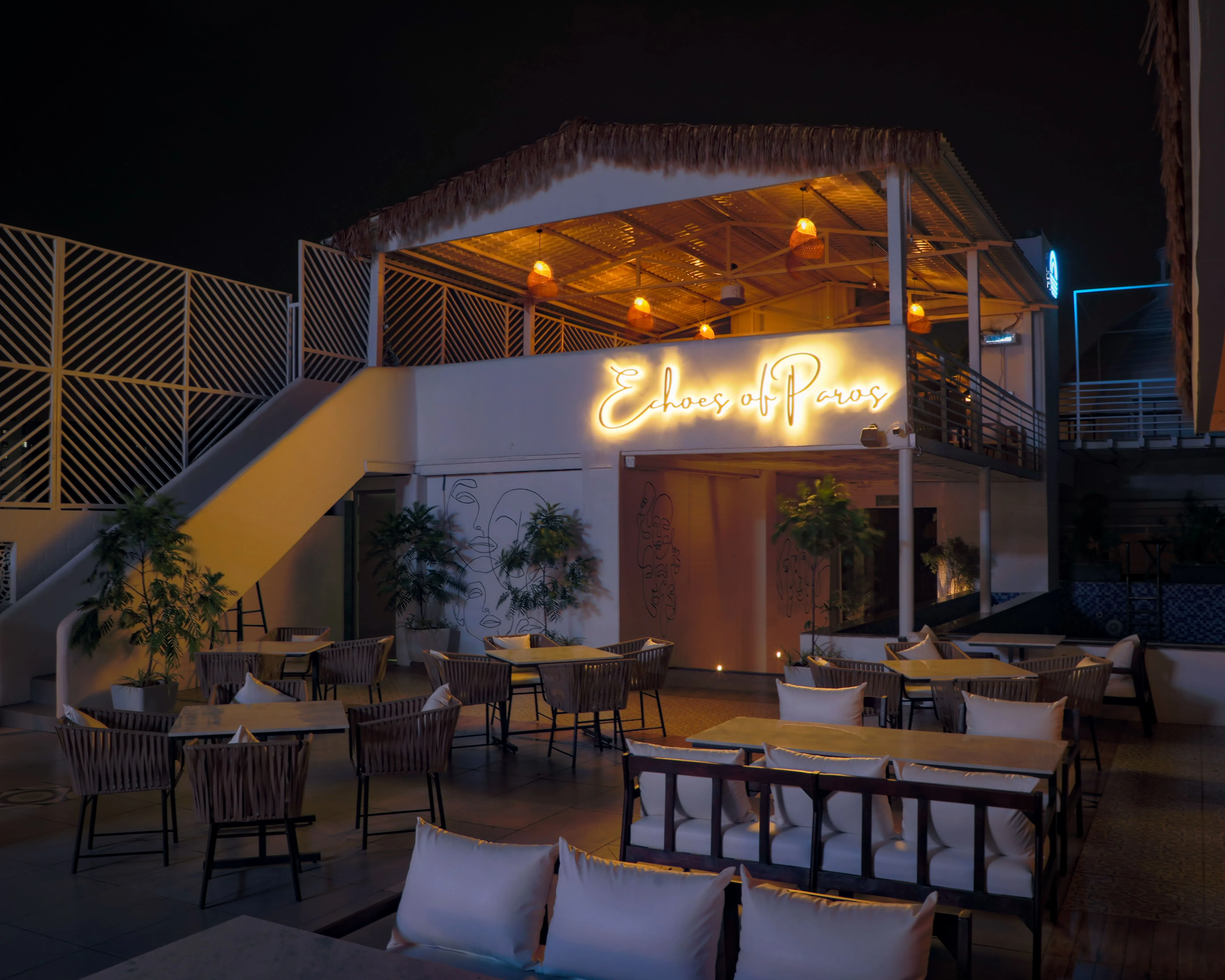 Outdoor seating area of a restaurant at night with tables, chairs, plants, and a lit sign reading 'Echoes of Paros'.