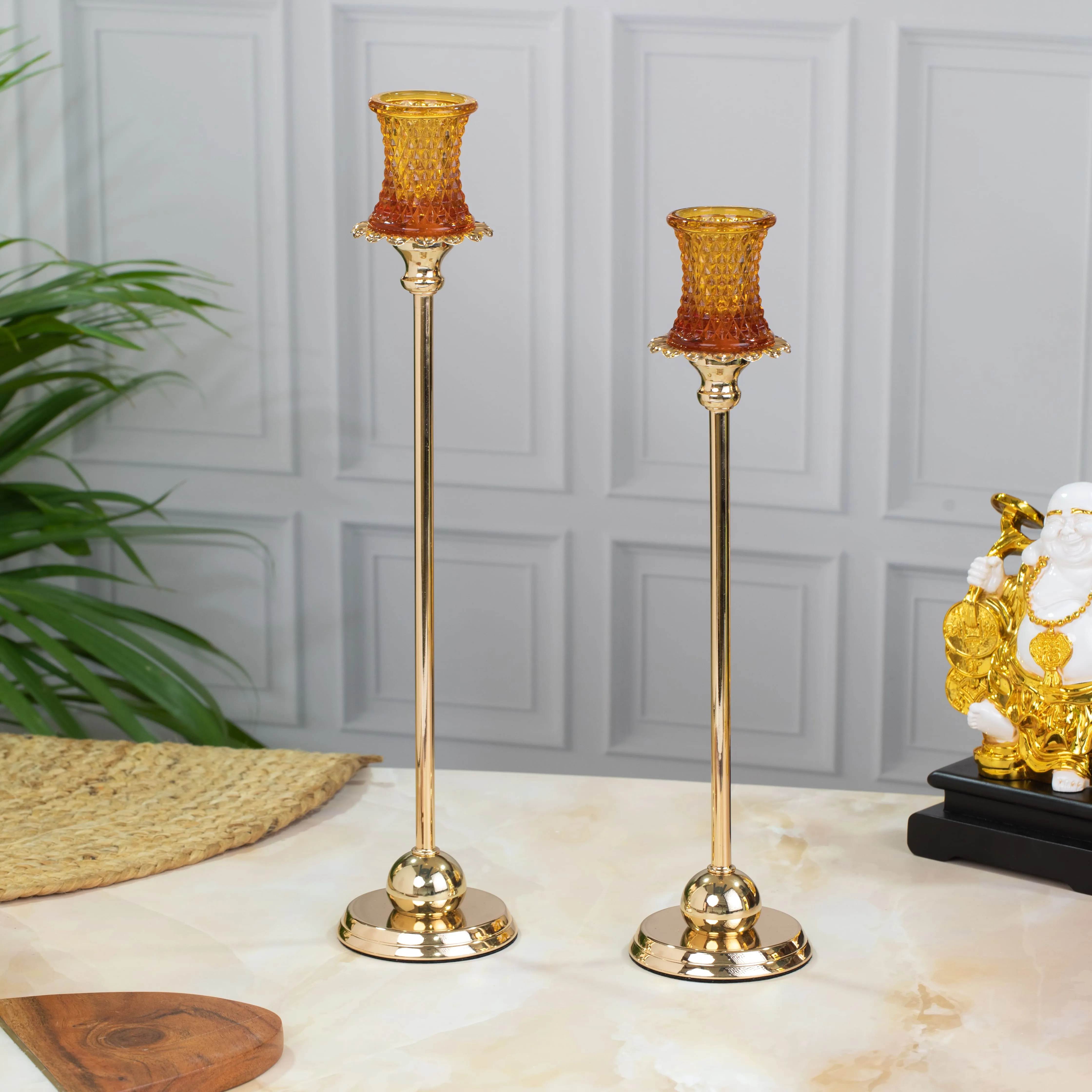 Two gold candlesticks with amber-colored textured glass candle holders on a marble surface, with a green plant and a golden statue in the background.