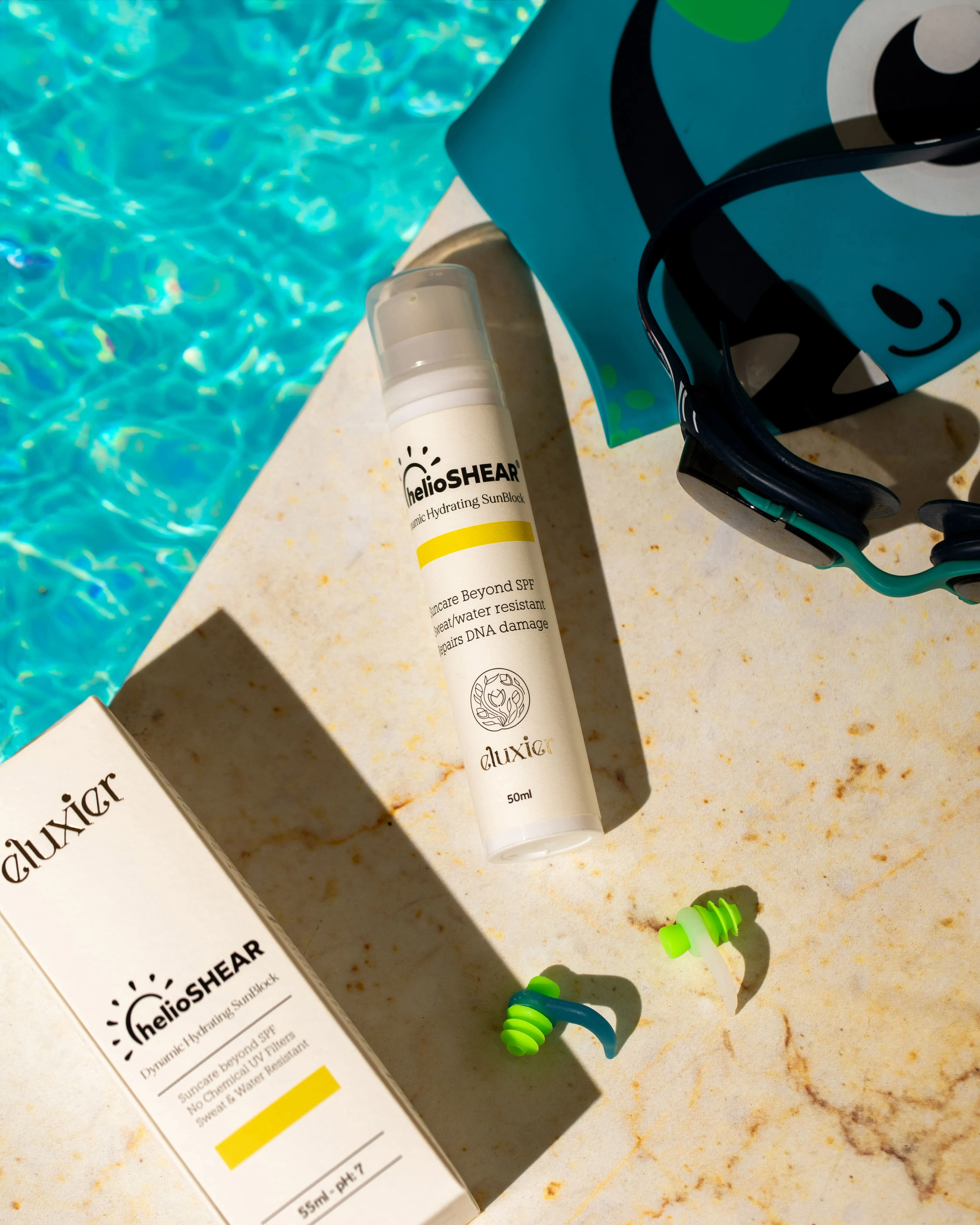 Tube and box of HelioSHEAR Dynamic Hydrating SunBlock on poolside marble, surrounded by swimming goggles and green earplugs.