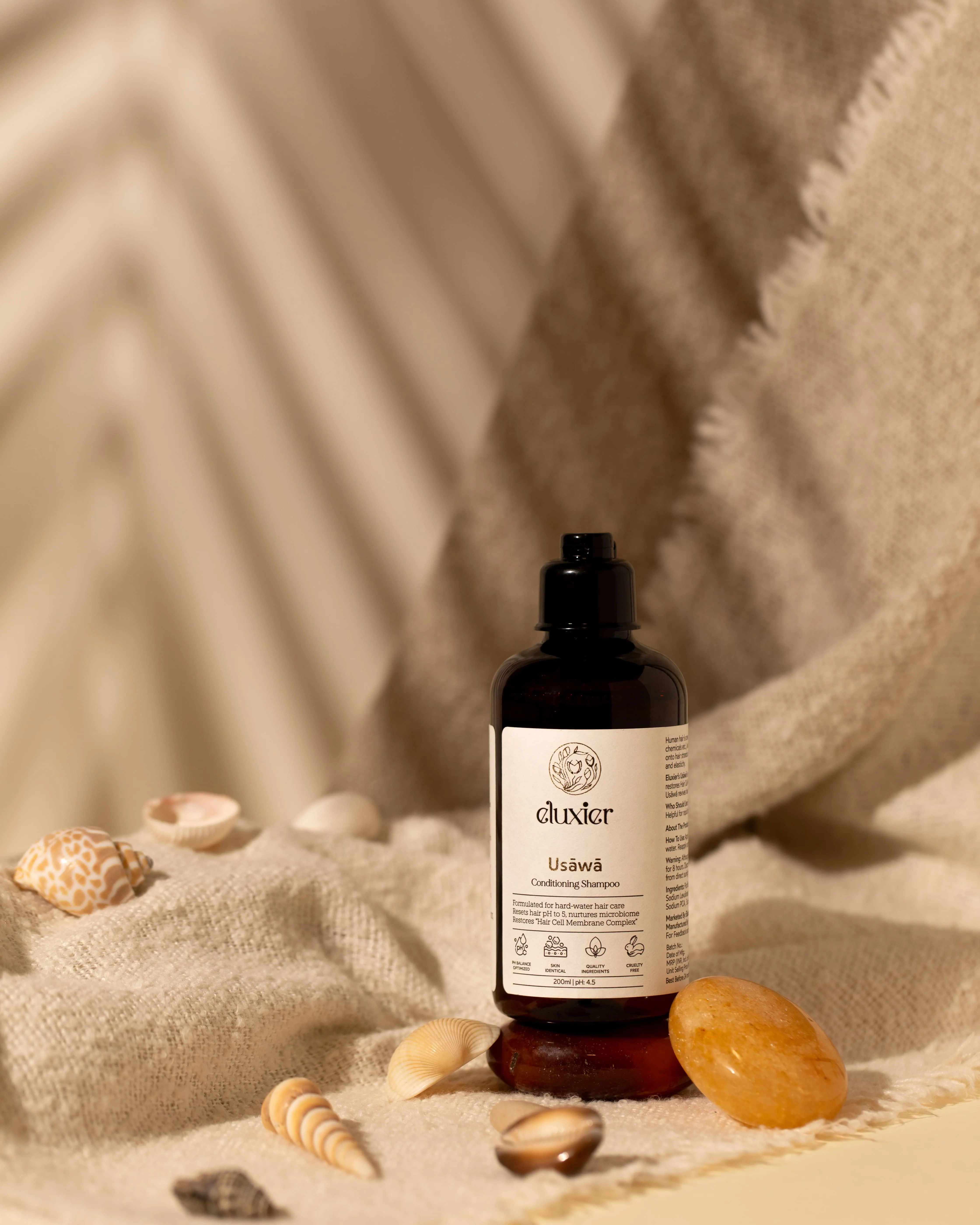 A dark bottle of Aluxier Usāwā Conditioning Shampoo placed on a beige textured fabric with seashells and a stone around it.