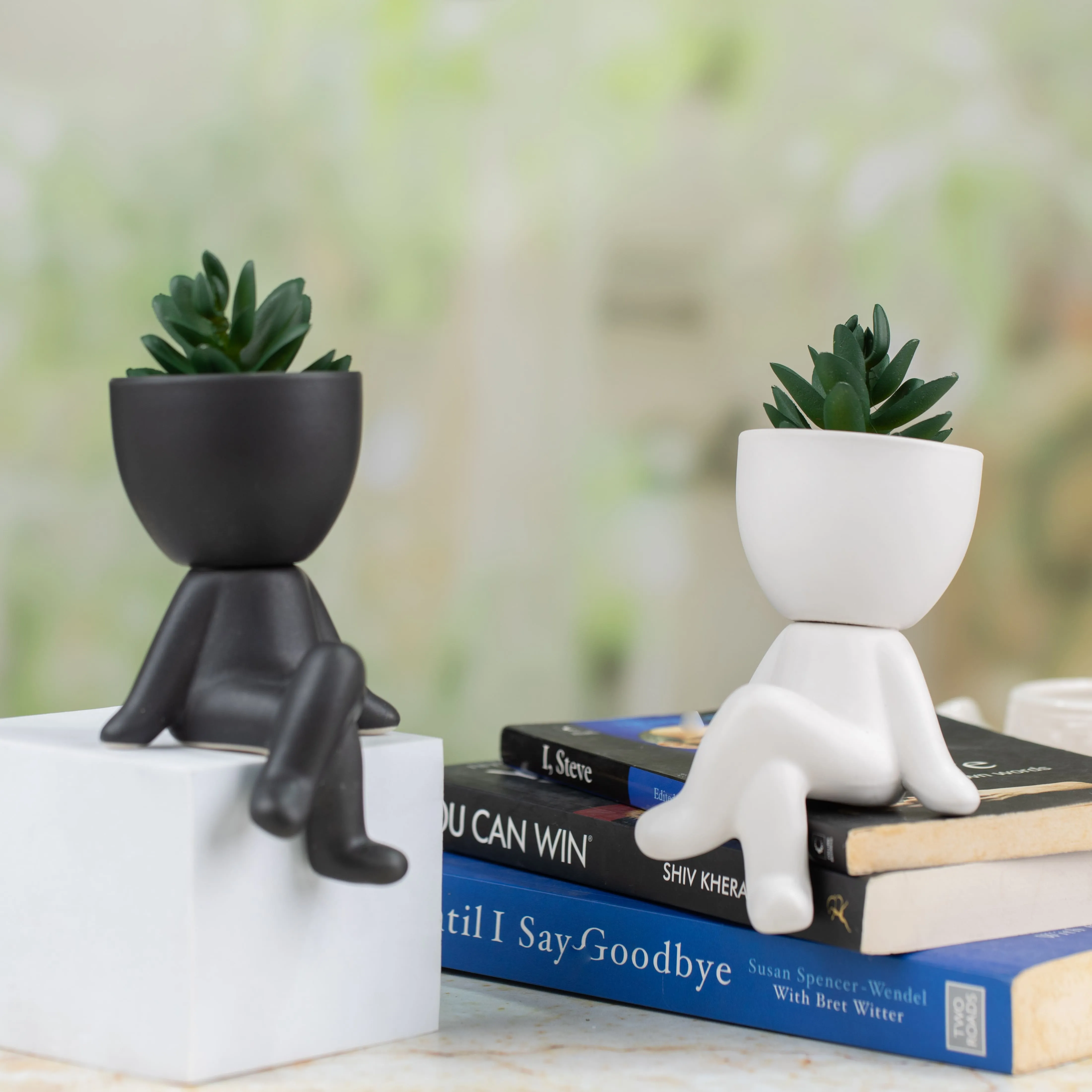 Black and white human-shaped succulent planters sitting on a white block and stacked books on a table.
