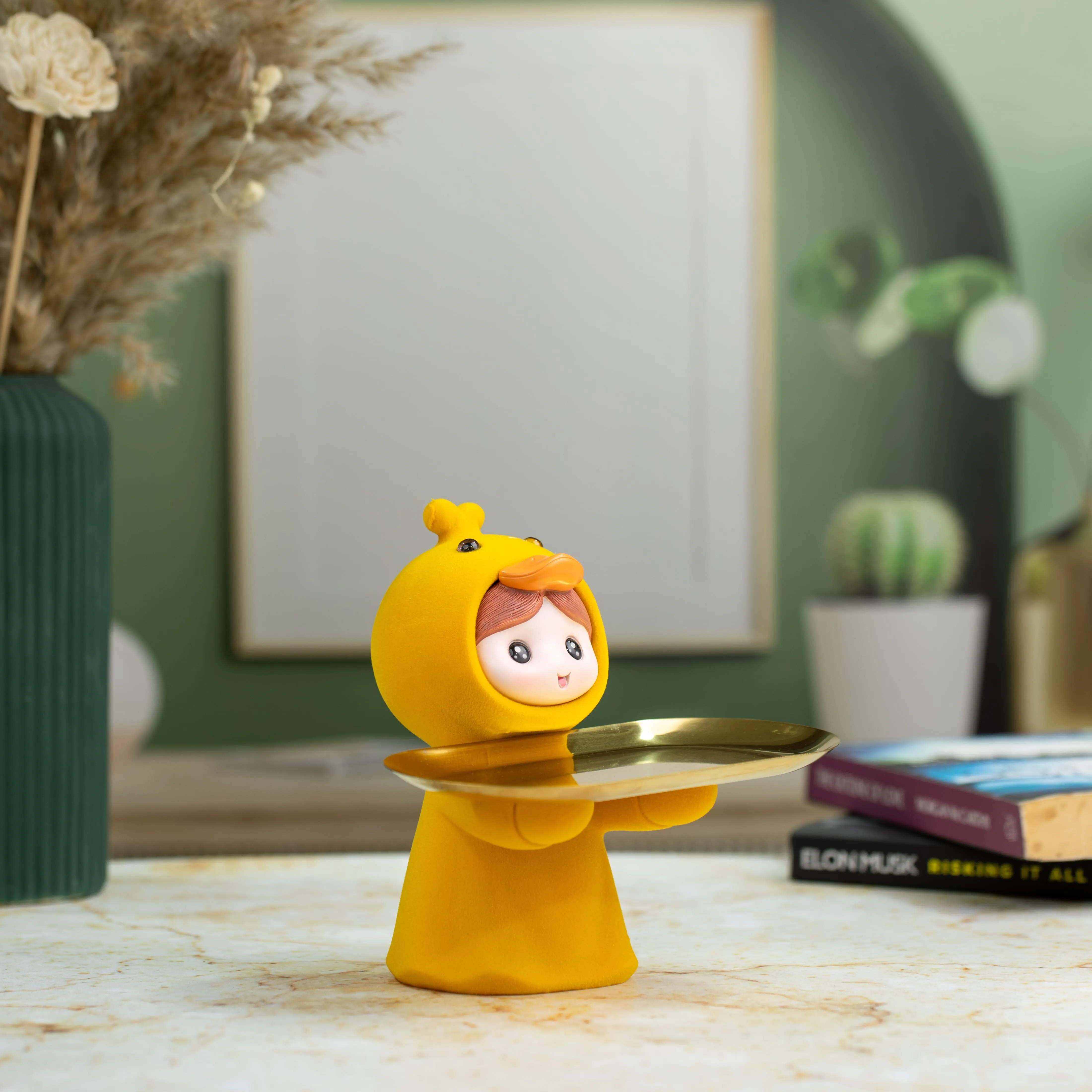 Small figurine wearing a yellow duck costume holding a golden tray on a marble surface with books and plants in the background.