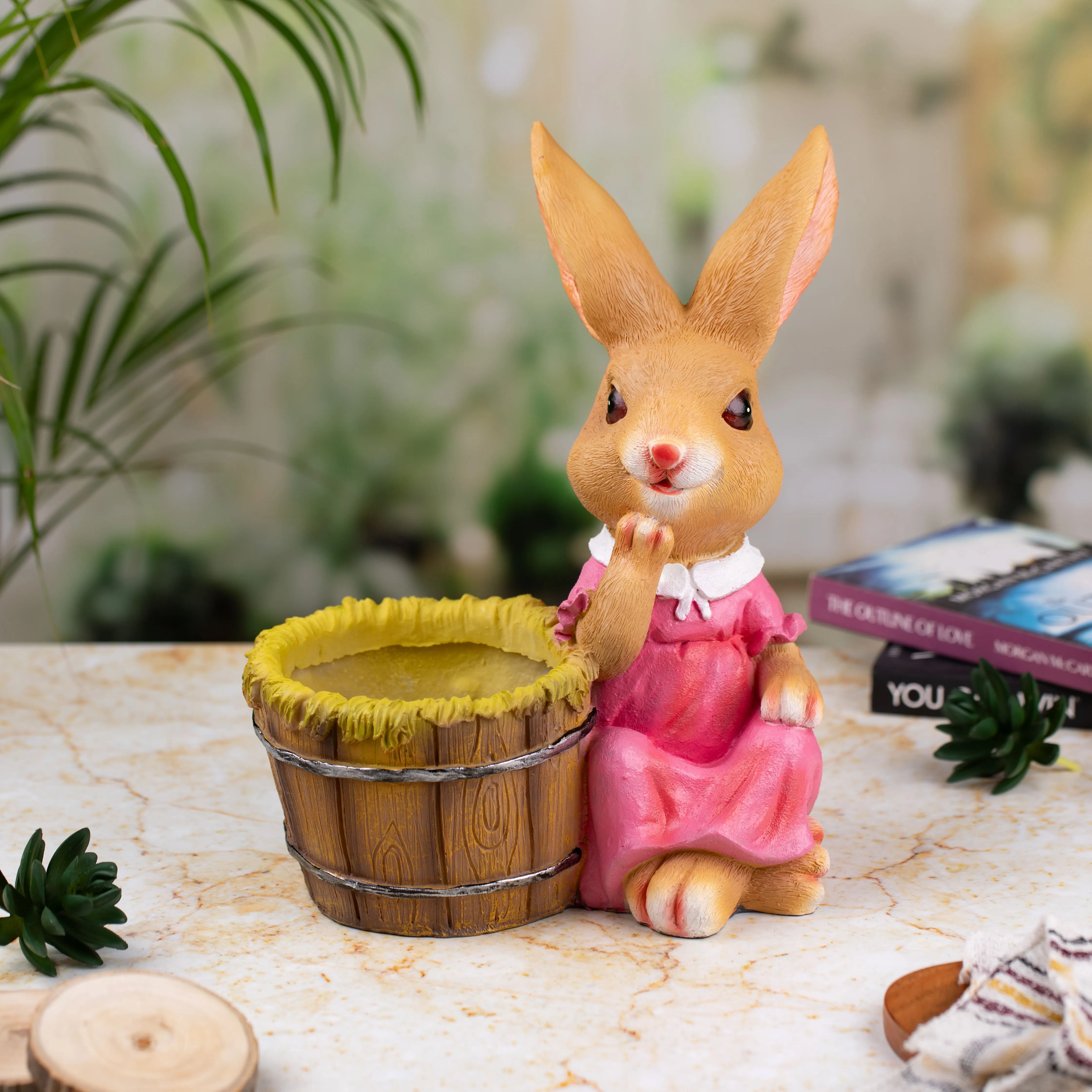 Ceramic bunny figurine in a pink dress sitting beside a wooden bucket with yellow trim on a marble surface with books and plants in the background.