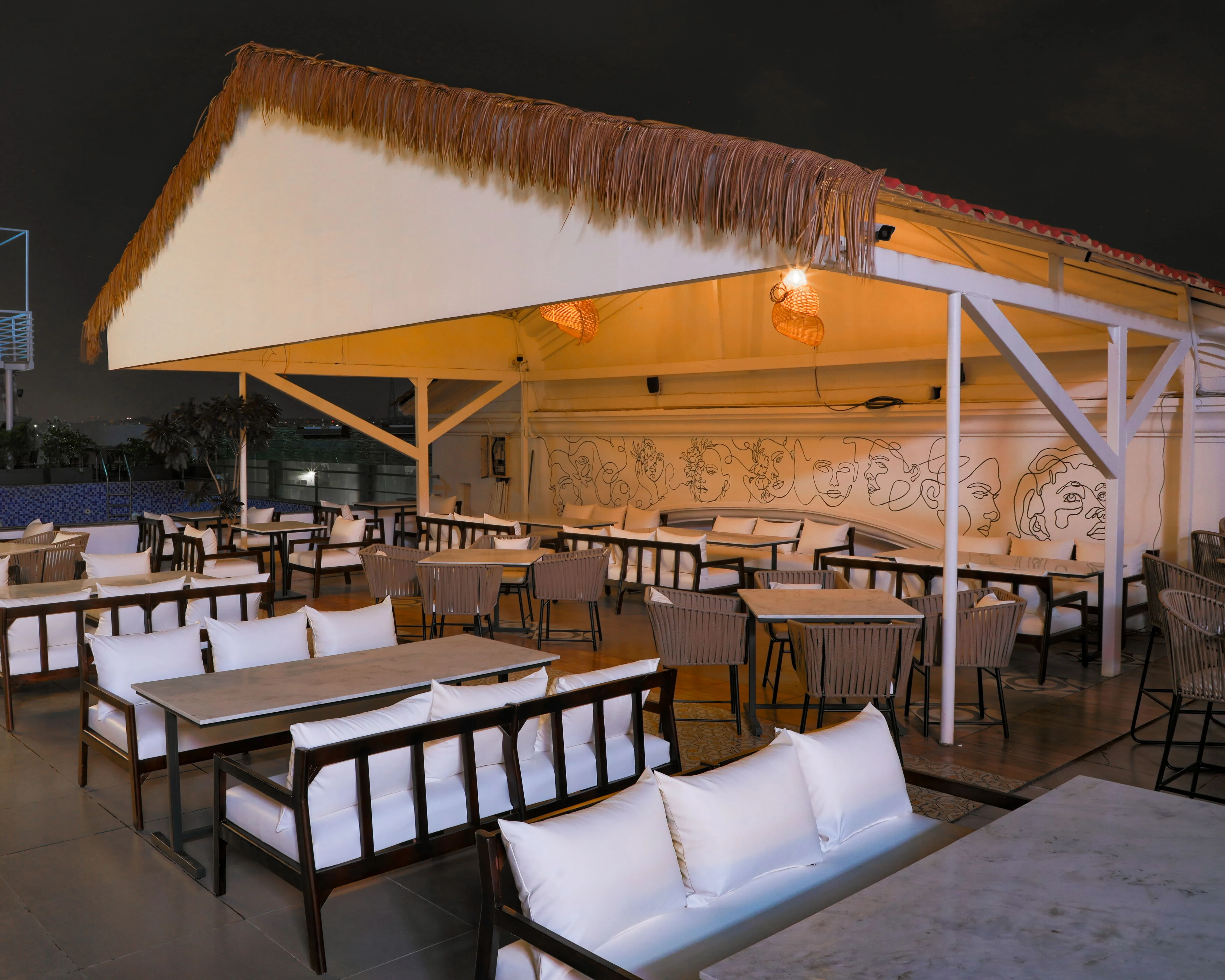 Outdoor restaurant seating area at night with cushioned wooden chairs, tables, and a thatched roof over part of the space.