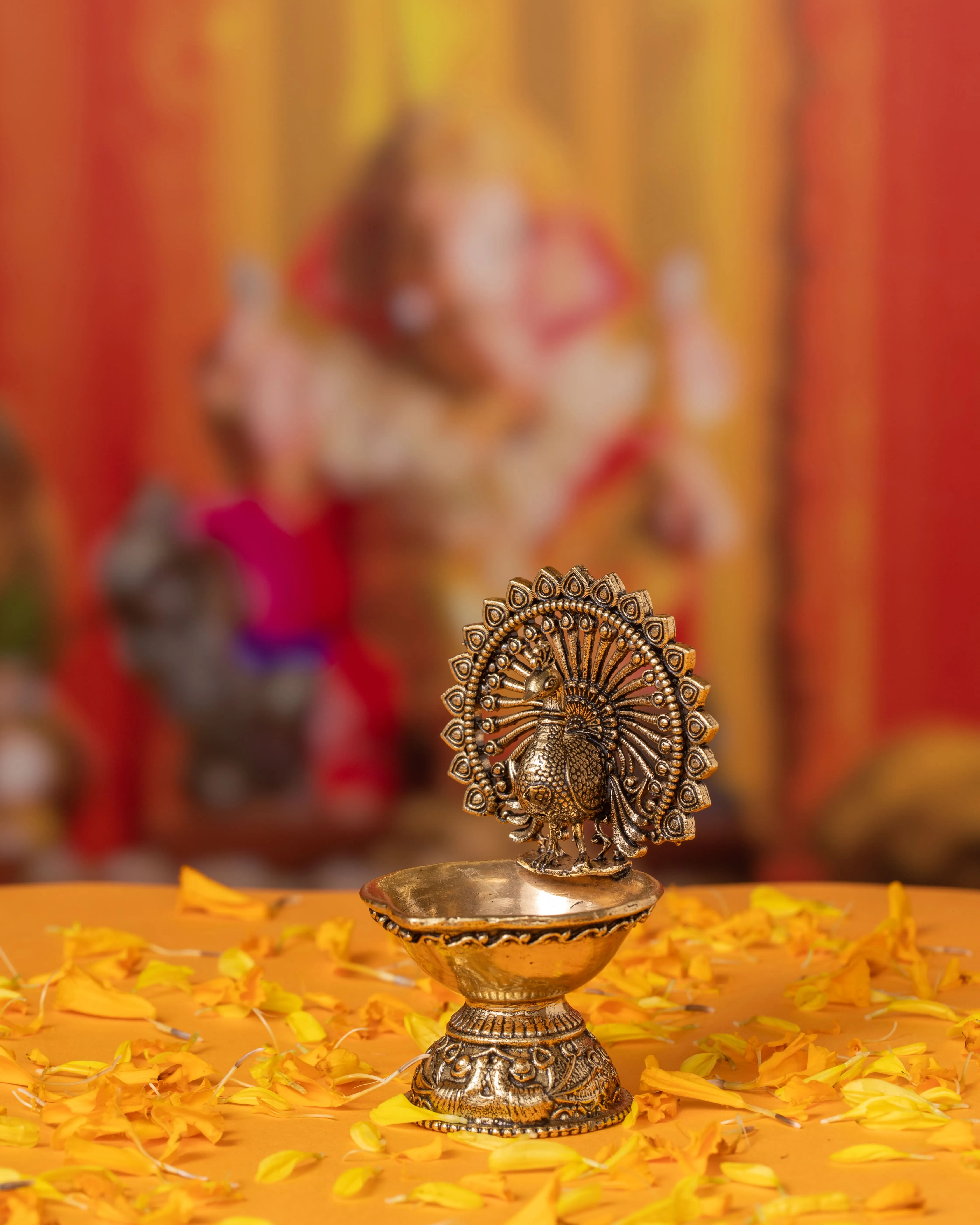 Decorative brass oil lamp with peacock design on a surface scattered with yellow flower petals and a blurred colorful background.