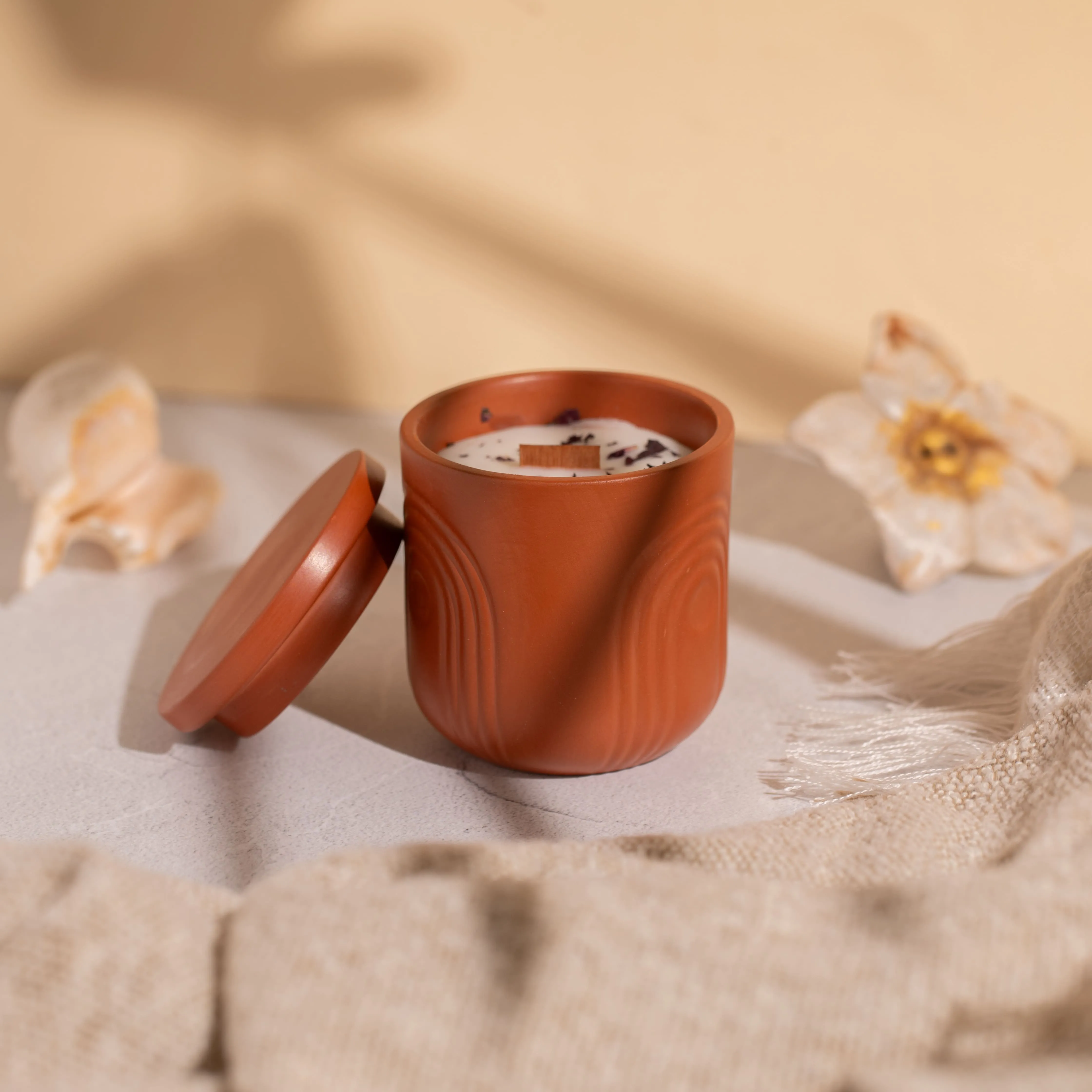 Terracotta candle jar with a wooden wick, lid leaning against it, placed on a surface with decorative seashells and a beige fabric.