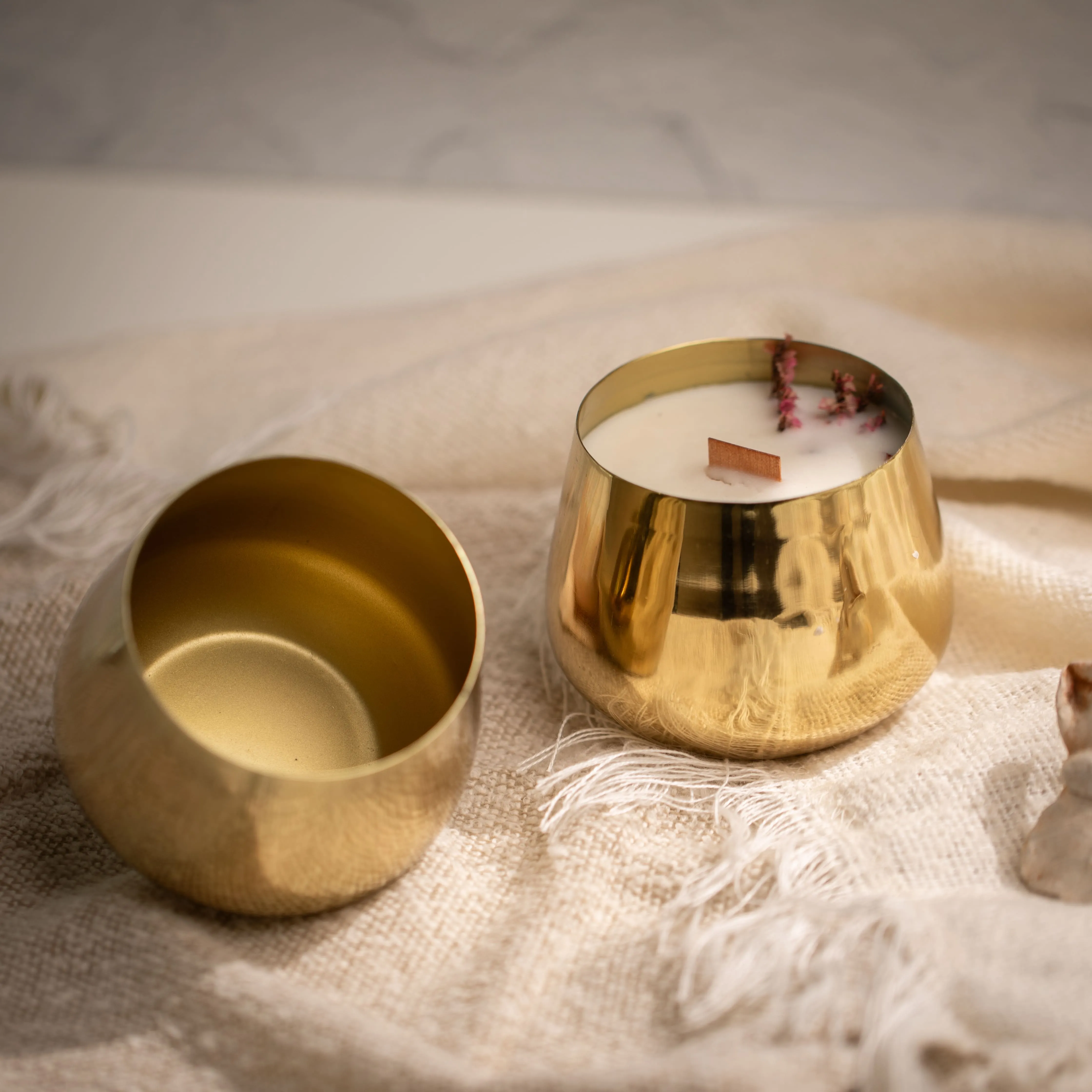 Golden metal container holding a white candle with a wooden wick and dried flowers, next to its empty matching lid on a beige textured fabric.