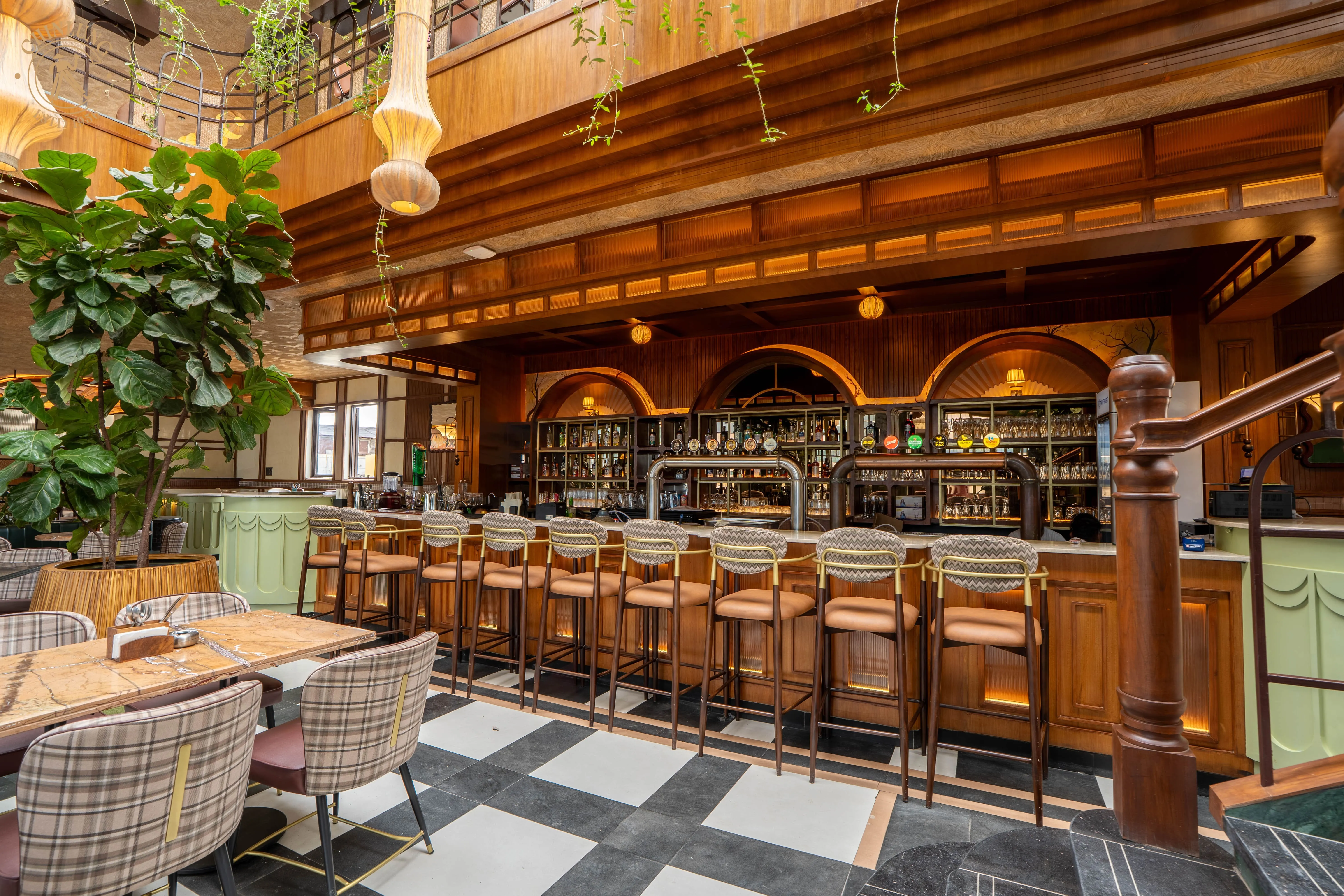 Stylish bar with wooden high chairs, checkered floor, green potted plant, and hanging pendant lights in a cozy indoor setting.