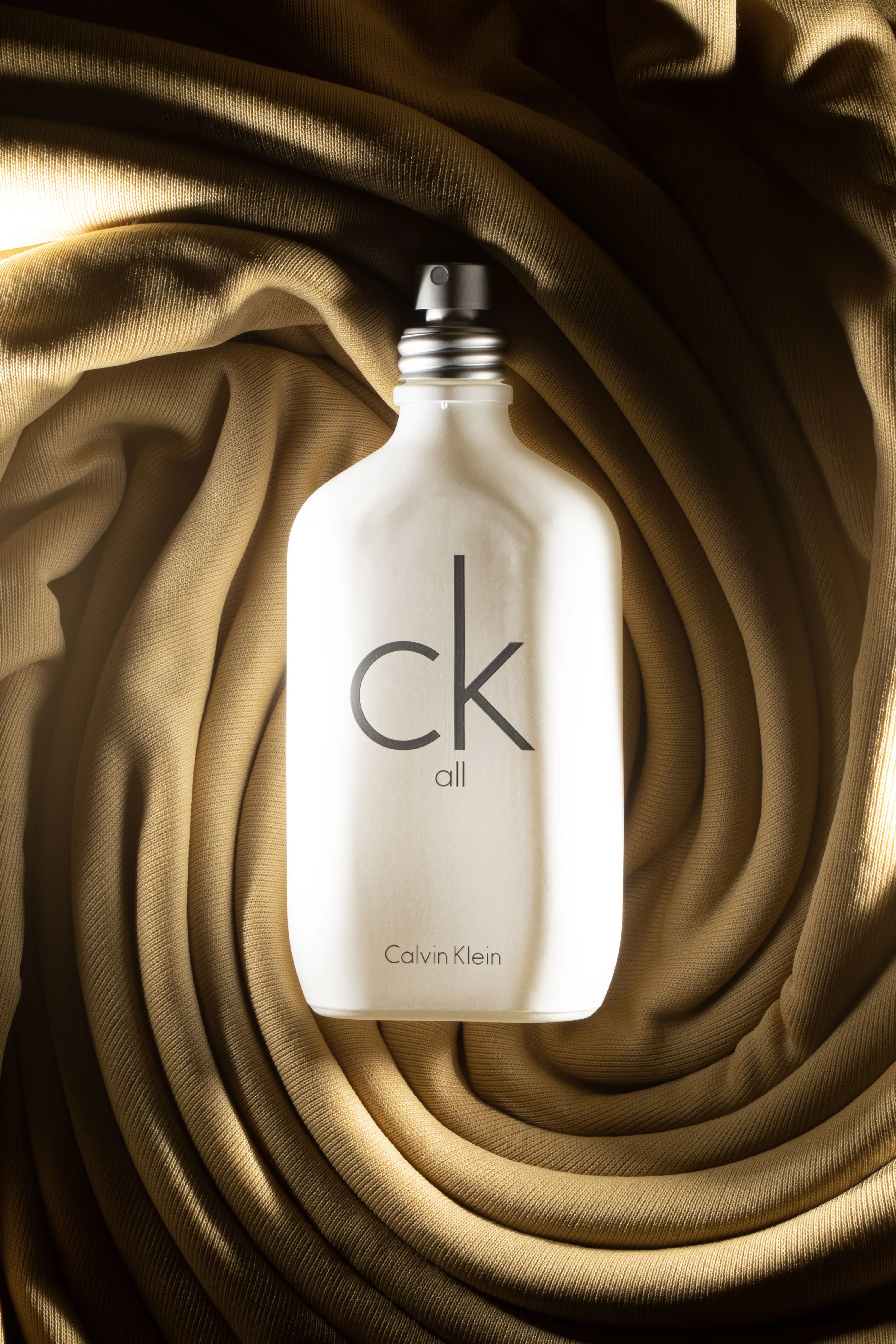 White Calvin Klein CK All perfume bottle on a textured, folded beige fabric background.