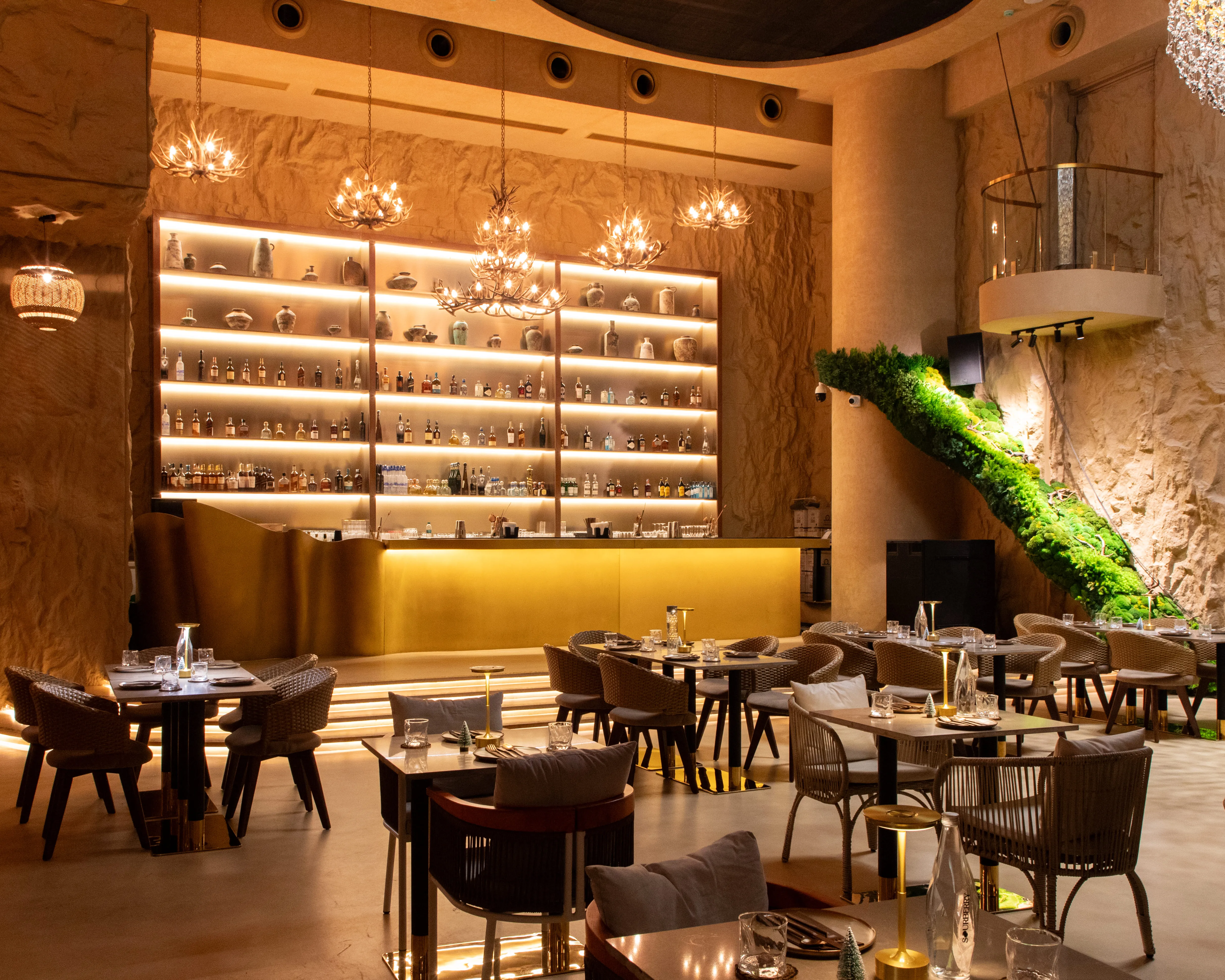 Modern restaurant interior with lit bar shelves, antler chandeliers, and tables set with glassware and candles.
