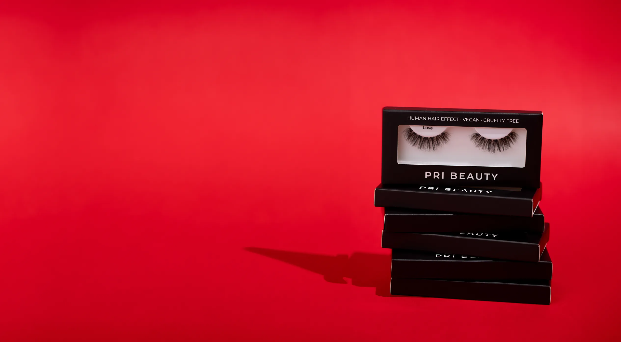 Stack of five black PRI BEAUTY eyelash boxes with one open showing false eyelashes labeled 'Love' on a red background.