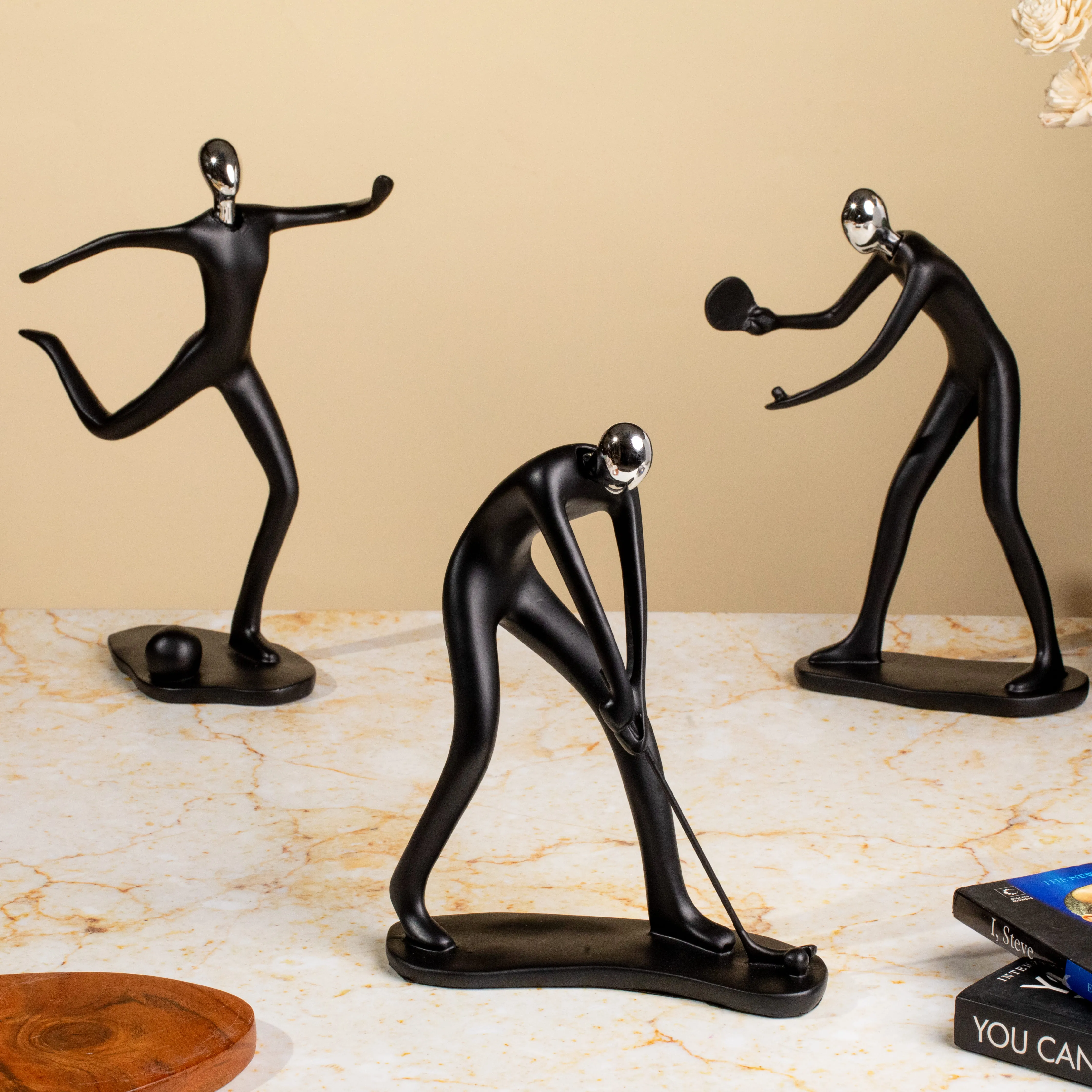 Three sleek black metal sculptures depicting athletes playing soccer, golf, and table tennis on a marble surface.