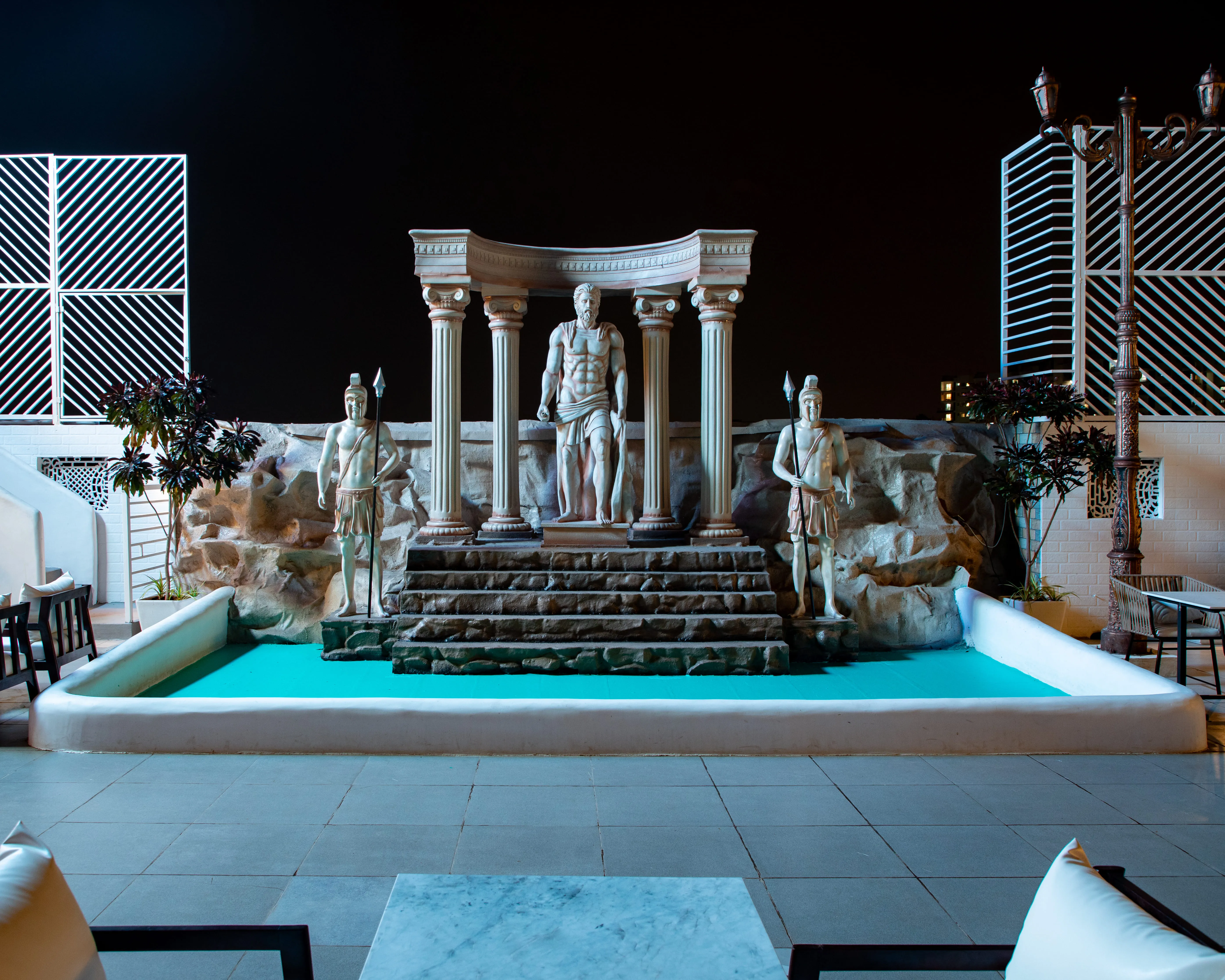 Night view of a classical Greek-style statue of a muscular man standing under a columned arch, flanked by two warrior statues with spears, in front of a small blue pool.
