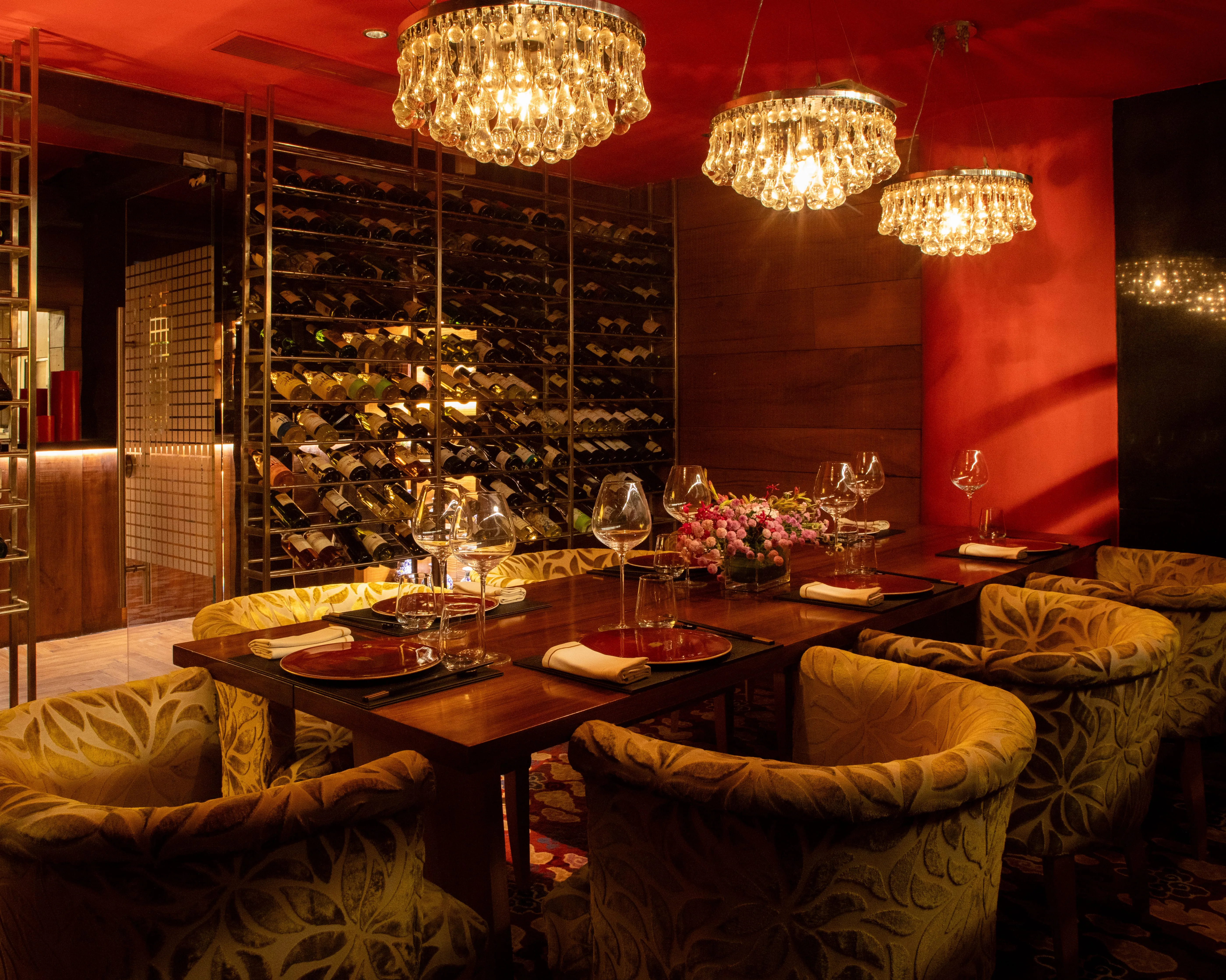 Elegant dining room with a wooden table set with plates, wine glasses, and floral centerpiece, surrounded by upholstered chairs and wine racks on the wall.