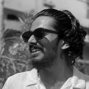 Man with a beard and mustache wearing sunglasses and a white shirt, smiling and looking to the side outdoors.