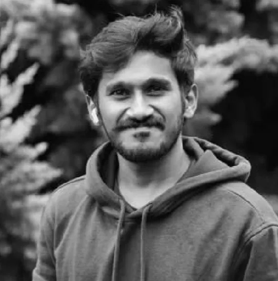 Young man with a beard and mustache wearing a hoodie, smiling slightly, with blurred foliage background.