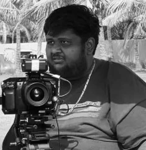 Man sitting behind a professional camera mounted on a tripod with palm trees in the background.