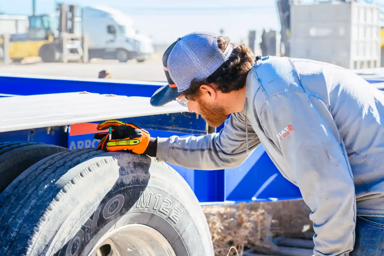 Technician conducting a DOT inspection on a commercial trailer, closely examining tire condition and tread depth in a well-lit outdoor yard.