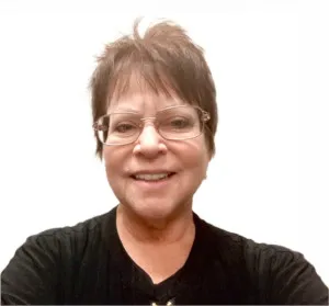 Smiling woman with short brown hair wearing glasses and a black top against a white background.