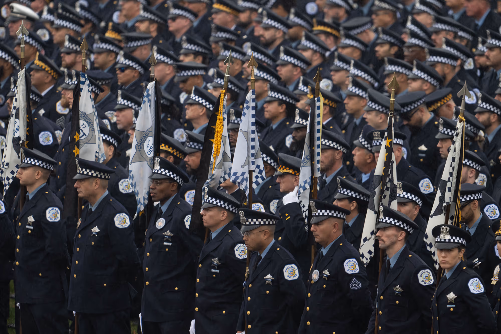 Rows of Chicago police officers in uniform standing solemnly, holding flags during a formal event.
