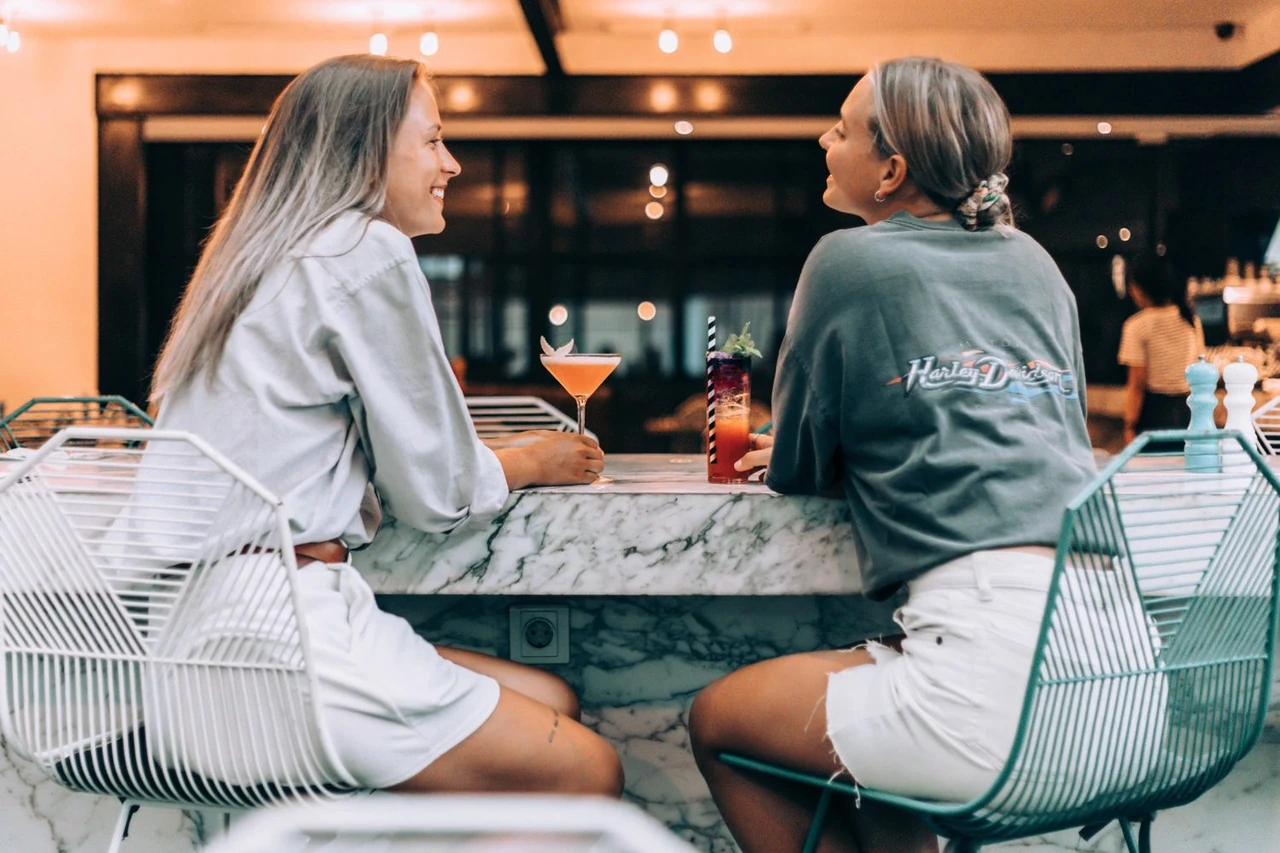 Two smiling women chat at a white marble bar with colorful cocktails. One wears a Harley Davidson shirt.