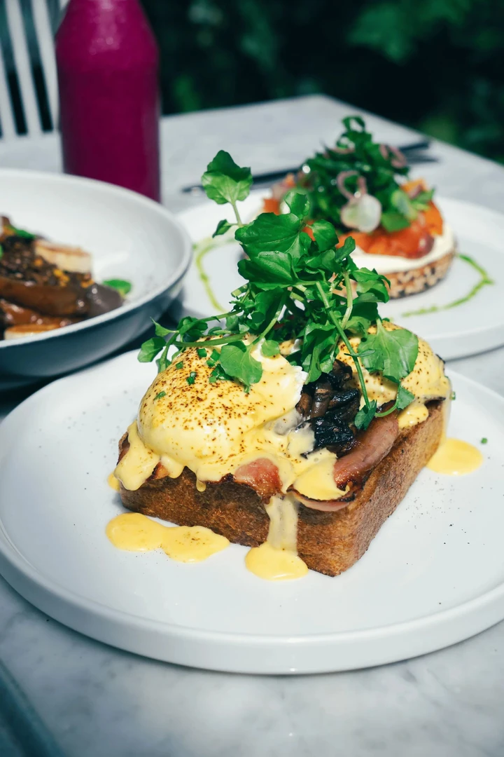 Eggs Benedict: poached eggs, bacon, mushrooms, hollandaise dripping on thick toast, topped with watercress. Other plates blurred.
