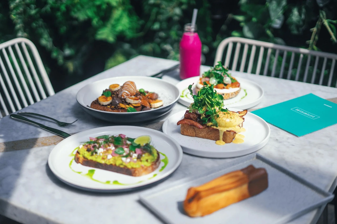 Vibrant brunch table outdoors with plates of avocado toast, eggs benedict, French toast, a pink smoothie, and a menu.