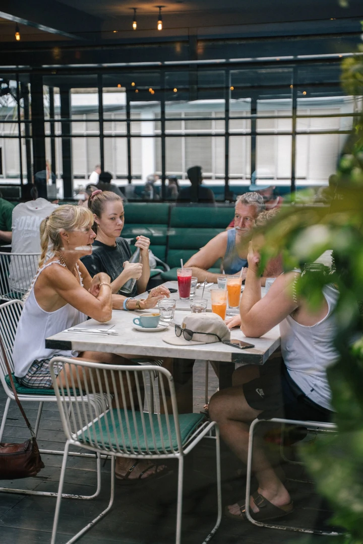Four people around a cafe table with coffee and colorful juices. White chairs, green cushions, and large windows create a bright scene.